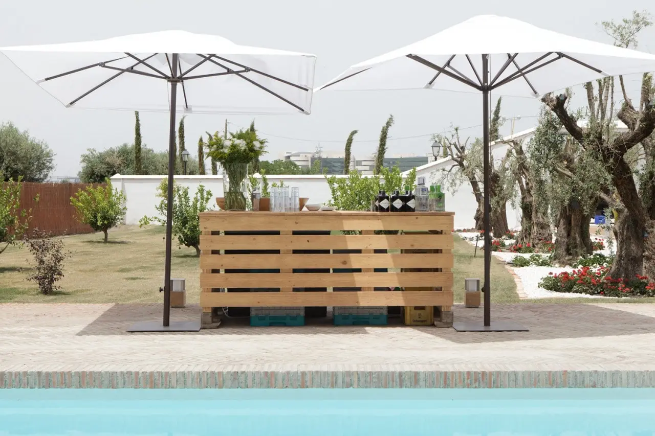 Una barra de madera junto a la piscina, bajo sombrillas blancas, con olivos detrás