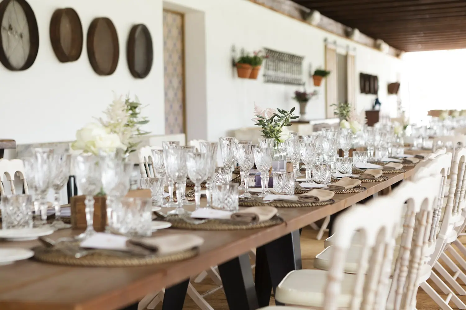 Una mesa larga puesta bajo el porche, con copas, flores y bajoplatos de esparto