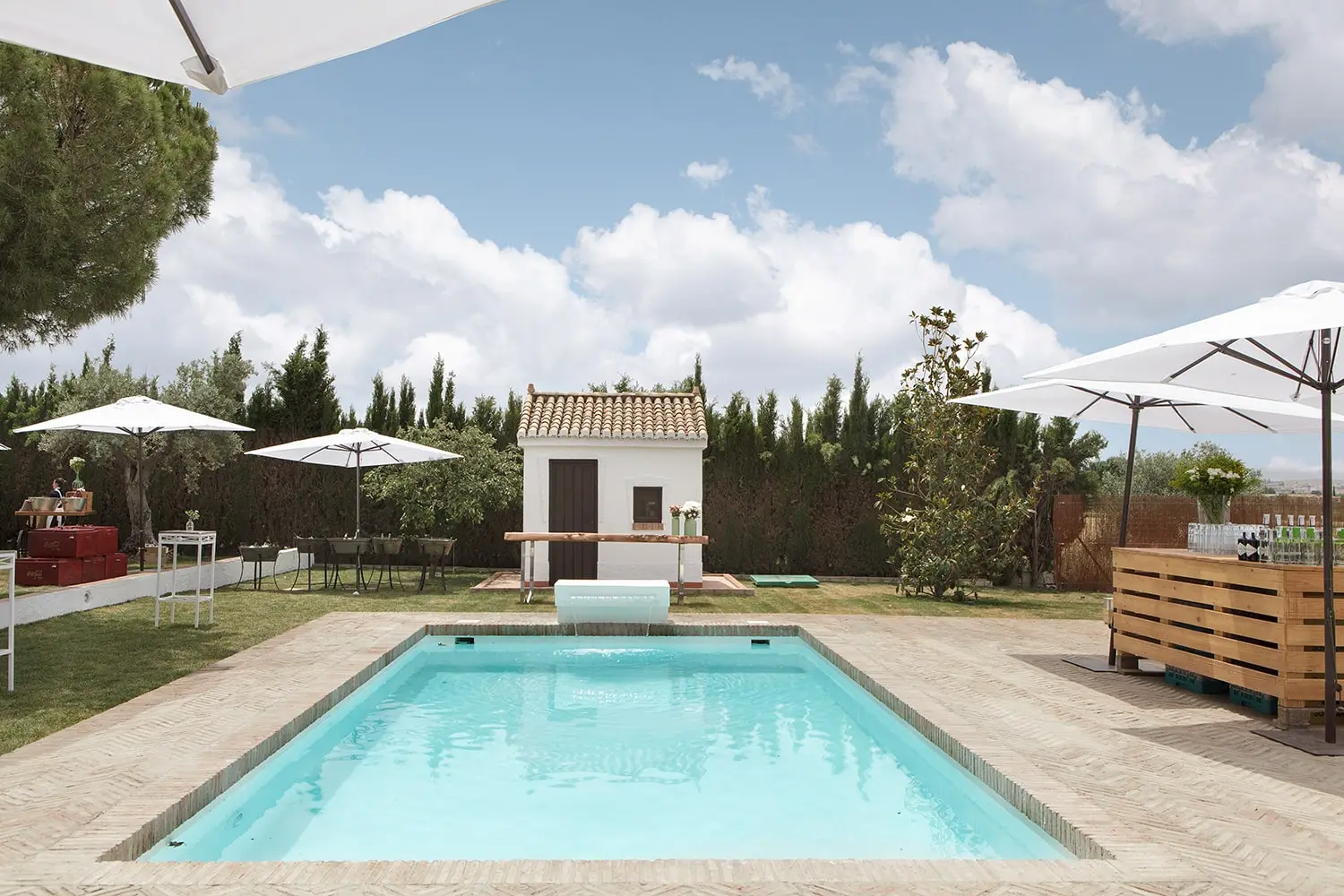 La piscina entre el césped y las sombrillas, con una casita blanca al fondo