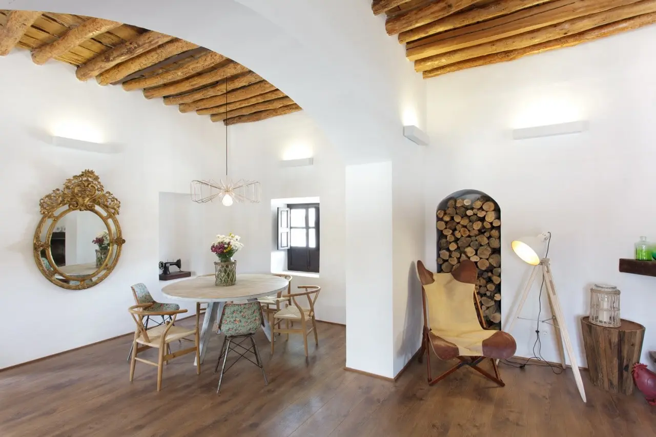 El salón con chimenea: vigas de madera, un espejo dorado, leña apilada y una mesa redonda